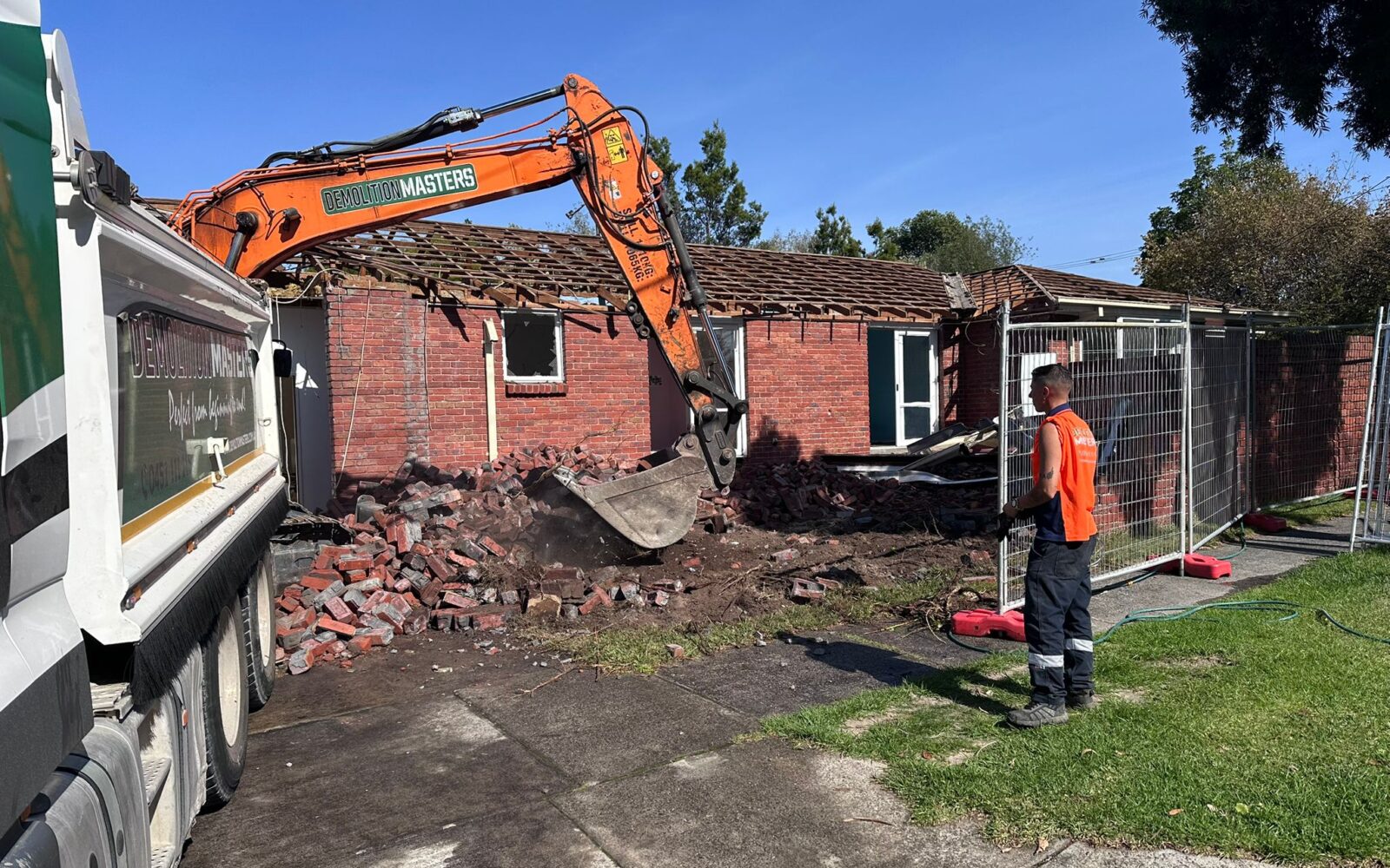 Branded Demolition Masters orange excavator demolishing a single-storey brick home