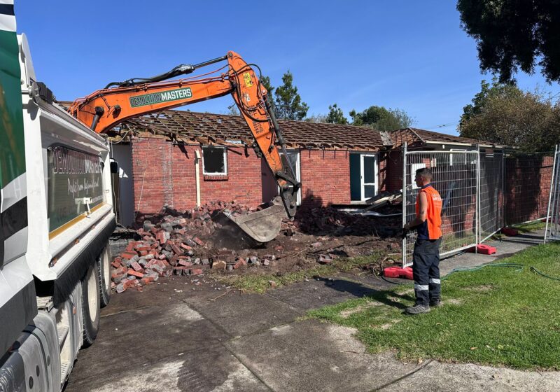 Branded Demolition Masters orange excavator demolishing a single-storey brick home