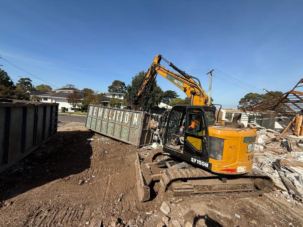 Two Demolition Masters branded bins side by side on a demolition site with an excavator loading