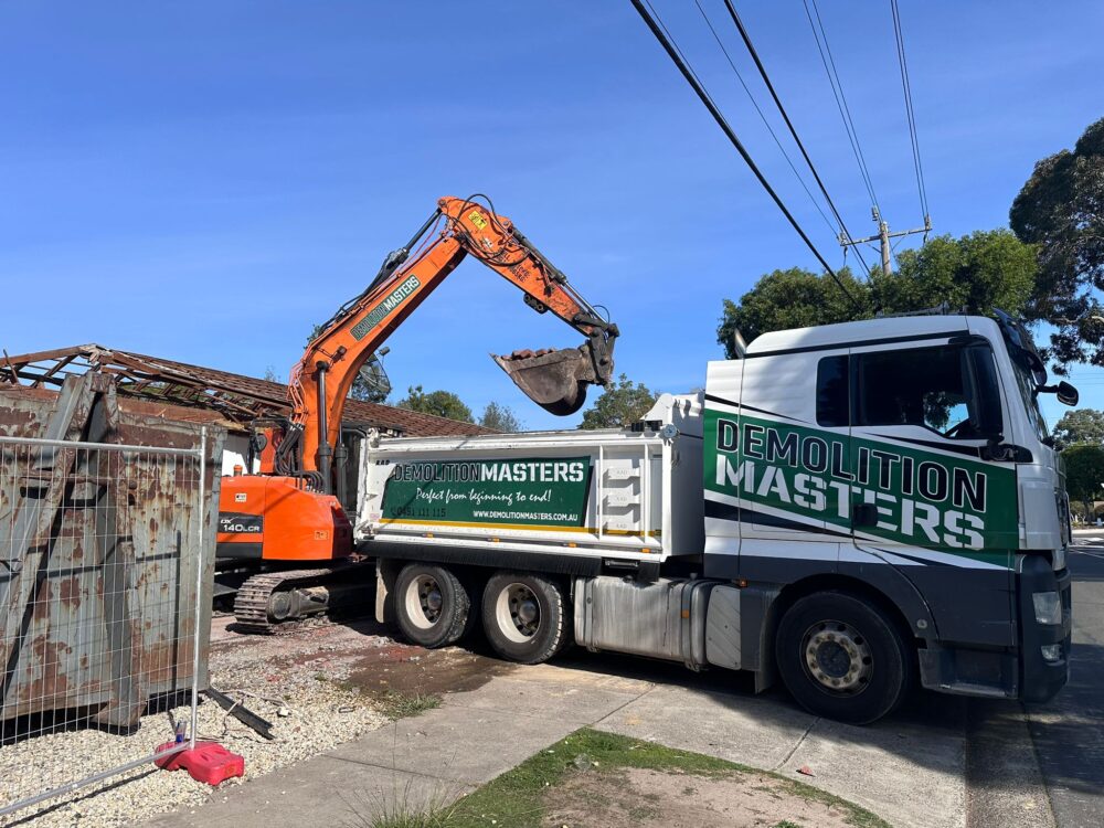 Branded Demolition Masters orange excavator loading dirt into a branded green and white truck