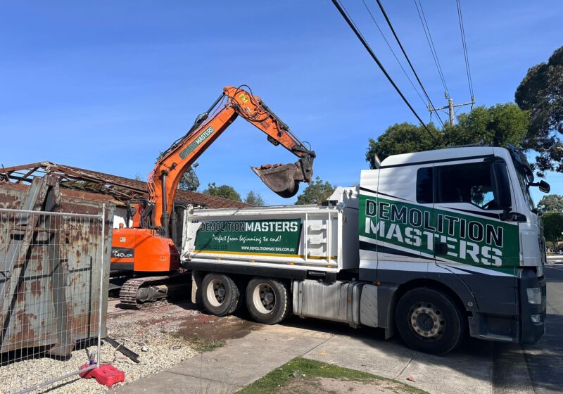 Branded Demolition Masters orange excavator loading dirt into a branded green and white truck