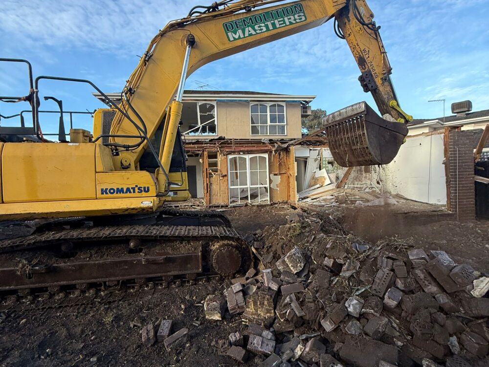 Yellow Komatsu excavator with Demolition Masters branding tearing into a brick home