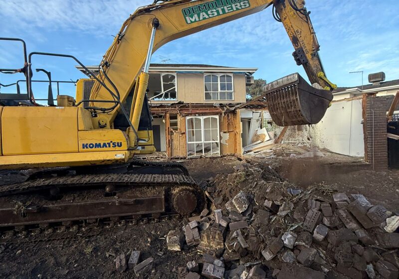 Yellow Komatsu excavator with Demolition Masters branding tearing into a brick home