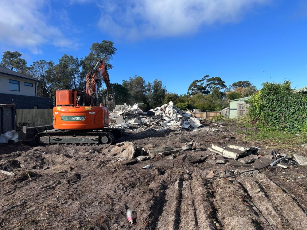 Branded Demolition Masters orange excavator beside a cleared site with debris pile after demolition